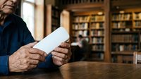 Senior man with wrinkled hands holding blank white tablet device at wooden table in library with bookshelves, wearing blue shirt. Suitable for technology adoption, elderly learning, digital literacy.
