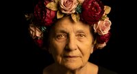 Elderly Caucasian woman wearing colorful floral crown with roses and peonies against dark background for portrait photography and artistic projects.