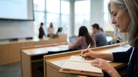 Young blonde woman student taking notes during university lecture in modern classroom with blurred classmates in background.