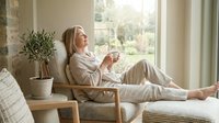 Middle-aged woman with blonde hair relaxing in wooden chair by window, holding coffee cup, wearing casual beige clothing, enjoying peaceful morning at home with natural sunlight and potted plant nearby.
