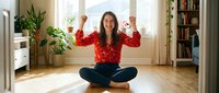 Young Asian woman celebrating success sitting cross-legged on wooden floor in bright modern living room with plants and bookshelves in background.