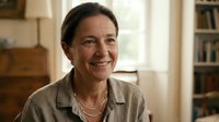 Smiling mature Caucasian woman in casual shirt at home office with warm lighting and bookshelves in background for lifestyle and professional content.