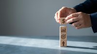 Businessman hands stacking wooden blocks with financial growth symbols including dollar sign, chart, target and arrow for business strategy concept.