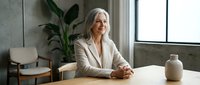 Mature woman with long white hair and fair complexion, wearing a beige blazer, seated at a wooden table in a modern office or home workspace with potted plants and natural light from large windows.