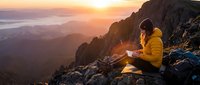 Woman in yellow jacket sitting on mountain rocks at sunrise, holding glowing lantern overlooking misty valley landscape. Adventure, travel, nature exploration.
