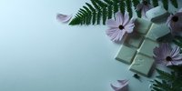 Pieces of white chocolate arranged with delicate purple daisy flowers and fern leaves on mint green background, creating elegant composition for confectionery design.