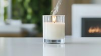 Burning white scented candle in glass holder with rising smoke trail against blurred cozy interior background with fireplace, creating relaxing spa atmosphere.