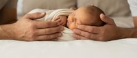 Newborn baby sleeping peacefully in protective father's hands on soft white bedding. Tender parenting moment showing care, love and family bonding at home.