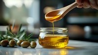 Golden honey dripping from wooden spoon into glass jar with fresh olives on kitchen counter for cooking and natural food preparation.