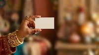 Closeup of woman's hand wearing gold bangles holding blank white card against blurred indoor background, suitable for business cards, invitations, and promotional designs.