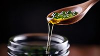 Golden olive oil dripping from wooden spoon with fresh herbs into glass jar on dark background, macro food photography with selective focus.