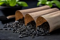 Black sunflower seeds spilling from brown paper rolls on dark background with green plants, dramatic lighting creates contrast for food photography and packaging.
