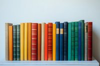Row of vintage hardcover books arranged by color on white shelf against light wall, featuring yellow, red, blue and green spines creating rainbow effect.