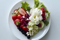 Fresh fruit and vegetable salad with strawberries, blackberries, cucumber, lettuce, and white flowers on round white plate against light background.