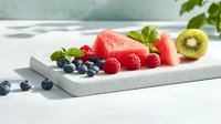 Fresh summer fruits on white stone board - juicy watermelon slices, blueberries, raspberries and kiwi garnished with mint leaves in bright natural lighting.