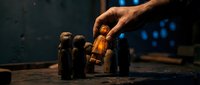 Hand selecting amber glass bottle from row of essential oil containers on wooden surface in warm atmospheric lighting for aromatherapy and wellness applications.