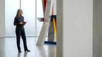 Middle-aged Caucasian woman in professional business attire presenting or discussing modern abstract art installation in contemporary gallery space with minimalist decor.