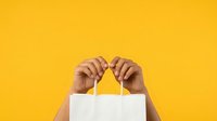 Person with dark skin tone holding white shopping bag against bright yellow background, representing retail, consumer lifestyle, and commerce.