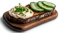 Open sandwich on dark rye bread topped with creamy spread, fresh cucumber slices and chopped herbs served on wooden cutting board against white background.
