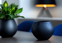 Modern black geometric vase with textured diamond pattern on table with green plant and blue chairs in soft focus background, contemporary home decor styling.