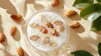 Fresh raw almonds and green leaves scattered around glass jar filled with almond milk on white background with natural sunlight and shadows.