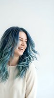 Happy young woman with vibrant blue teal hair laughing joyfully in casual white sweater against neutral background for lifestyle content.