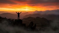 Silhouette of person with raised arms celebrating success on mountain peak during golden sunrise over layered mountain ranges with dramatic orange sky.