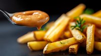 Golden french fries with fresh rosemary and dripping sauce on spoon against dark background, close up view of crispy potato strips with herbs and dip.