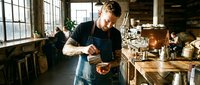 Professional barista preparing specialty coffee in rustic industrial cafe with vintage equipment and natural lighting from large windows.