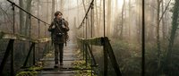 Man in military uniform standing on wooden suspension bridge in misty forest with bare trees and fog.