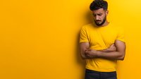 Middle Eastern man with beard wearing yellow t-shirt showing negative emotion with arms crossed against vibrant yellow background, studio portrait with copy space.