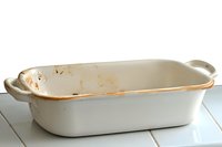 Empty white ceramic baking dish with handles shows signs of use and cooking stains on cream-colored wooden surface, rustic kitchen setting.
