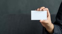 Businessman in dark suit holding blank white business card against gray background, ready for contact information or branding mockup.