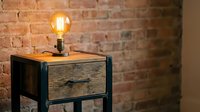 Vintage Edison bulb on industrial nightstand with wooden drawer against exposed brick wall, creating warm ambient lighting for home decor.