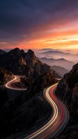 Winding mountain road with light trails at sunset showcasing dramatic rocky peaks and layered mountain ranges under colorful sky for travel and adventure concepts.