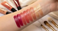 Woman hand with colorful lipstick swatches testing makeup shades from deep burgundy to nude pink tones with cosmetic brushes and tubes on marble surface.