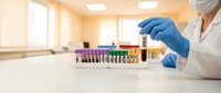 Lab technician in blue gloves holding blood sample tube with rack of colorful test tubes in medical laboratory background.