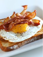 Crispy bacon strips and sunny side up egg on toasted bread slice with black pepper seasoning for breakfast meal preparation and cooking content.