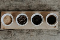 Coffee preparation stages on wooden board with raw sugar, roasted beans, ground coffee, and brewed coffee in white bowls.