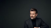 Middle-aged man with light skin tone, dark hair with gray streaks, and facial hair wearing a charcoal blazer and black turtleneck against a dark studio background for corporate and professional use.