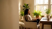 Young African American woman relaxing at home with laptop in cozy living room filled with green plants and natural sunlight streaming through windows.