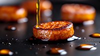 Seared foie gras medallion with caramelized surface and honey drizzle on dark background, dramatic lighting highlights golden drops and sea salt crystals.