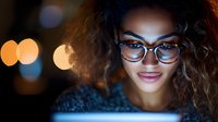 Young African American woman with curly hair and glasses illuminated by blue light from screen, smiling while working late at night with bokeh background.