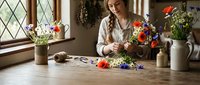 Young woman florist arranging colorful wildflowers in rustic kitchen with diamond-paned windows. Professional flower arrangement workspace scene.