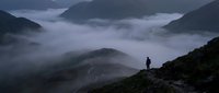 Silhouette of a person standing on a rocky mountain ridge overlooking a vast valley filled with dense fog and mist, surrounded by forested peaks at dawn or dusk.