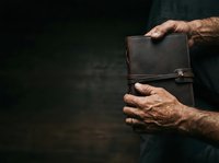 Mature man with medium skin tone holding worn leather journal or notebook, wearing dark clothing, studio shot with dark background for editorial and lifestyle use.
