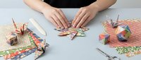 Woman crafting colorful origami paper cranes and geometric shapes on desk with decorative papers and scissors for creative hobby project.