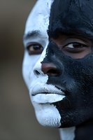 Face painted in contrasting black and white split design, close-up portrait showing dramatic makeup artistry and intense gaze against dark background.