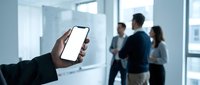 Hand holding blank white smartphone screen in modern office with blurred business team meeting in background for mobile app mockup and digital presentation.