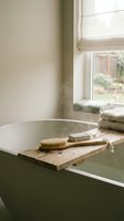 Rustic wooden bath caddy with natural bristle brush and folded towels across white bathtub in bright bathroom with window light for spa relaxation.